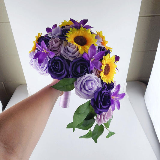 cascading Lilac, lavender, and dark purple real touch roses wedding bouquet. Lillies and sunflowers are scattered throughout. Handle is made up of satin lavender ribbon. 